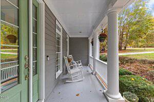A covered porch with a light green door, white columns, gray siding, two white rocking chairs, hanging flower baskets, and a view of a lawn with trees and shrubs.