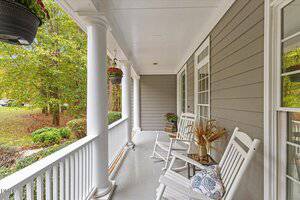 A covered porch with white columns and railing, gray siding, three white rocking chairs with cushions, potted plants, and a view of a green, wooded yard.