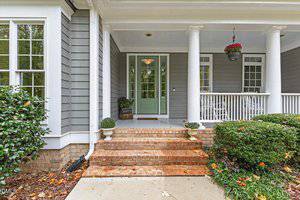 A brick front porch with steps leads to a light green door flanked by large windows. White columns support the roof, with hanging flower baskets and potted plants by the entrance. A white railing lines the porch.