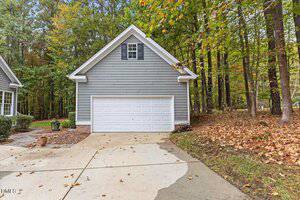 A gray detached two-car garage with a white door sits beside a wooded area. The garage has a gabled roof and a small upper window. Fallen leaves are scattered on the driveway and ground.