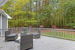 A gray wooden deck with a gray outdoor seating set and a small rug overlooks a wooded area with green trees. Potted flowers are placed along the deck railing, and the sky is partly cloudy.