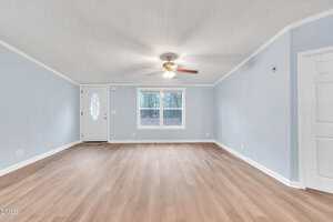 A bright, empty living room with light wood flooring, pale blue walls, a ceiling fan with lights, a white front door with an oval window, and two windows letting in natural light.