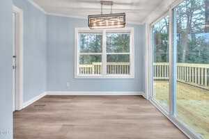 A small, empty room with light blue walls, wood-look flooring, a modern rectangular ceiling light, a window with a view of trees, and a large sliding glass door leading to a wooden deck outside.