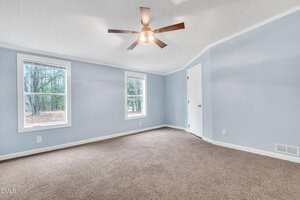 Empty room with light blue walls, two windows, brown carpet, a white door, and a ceiling fan with a light fixture; trees are visible outside the windows.