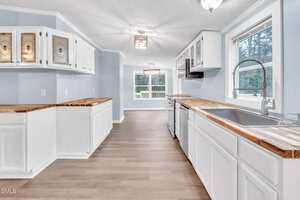 A spacious kitchen with white cabinets, butcher block countertops, a large sink, stainless steel dishwasher, light blue walls, and wood-look flooring. Windows provide natural light and offer a view of greenery outside.