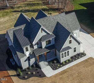 Aerial view of a modern, two-story house with gray siding, dark gray roofing, several gables, and black metal awnings. The property features a landscaped front yard and a wide concrete driveway.
