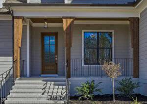 A front porch with gray steps leads to a wooden door with frosted glass; wooden pillars support the overhang. A black metal railing lines the porch, and small shrubs are planted in the mulched front yard.