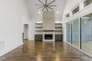 A modern, empty living room with hardwood floors, a stone fireplace with built-in shelves on both sides, a large ceiling fan, and sliding glass doors leading to an enclosed porch.