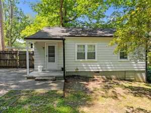 A small, single-story house with light-colored siding, a covered front porch, two windows, and a side door. The yard has patchy grass and is shaded by trees. A wooden fence runs along the left side.