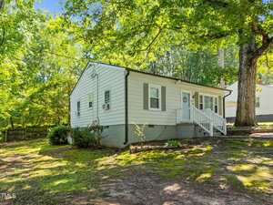 A small, single-story house with white siding, green shutters, and a front porch sits on a sloped lot shaded by large trees. Steps lead up to the porch, and the yard is mostly bare soil with some plants and grass.