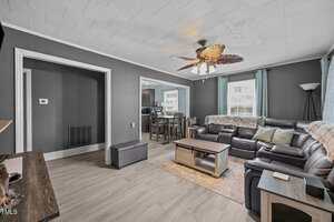 A living room with gray walls, light wood flooring, a sectional black leather sofa, a wooden coffee table, and a ceiling fan with leaf-shaped blades. A dining area and kitchen are visible in the background.
