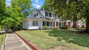A two-story white house with black shutters at 830 S Chestnut Street in Henderson features a covered front porch, red door, and large windows. It sits on a grassy corner lot with mature trees and a paved sidewalk along the edge.