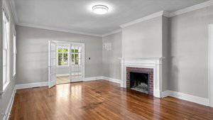 A living room with hardwood floors, light gray walls, a white fireplace with a brick surround, and double glass doors leading to a bright adjacent room with large windows.
