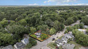 Aerial view of a residential neighborhood with a white house outlined in orange at a street corner, surrounded by trees and other homes; roads and greenery visible throughout the area.