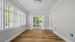 A bright, empty room with light gray walls, large windows on one side, wood flooring, and French doors opening to an outdoor area. White trim accents the windows, doors, and ceiling.