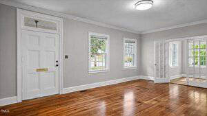 A bright, empty room with hardwood floors, light gray walls, a white front door, three windows, crown molding, and glass French doors leading to another room. A round ceiling light fixture is visible.