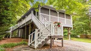 A raised house with a white staircase and railing, screened porch, and hanging flower pot. The home is surrounded by trees and greenery, with a gravel driveway and stepping stones leading to the stairs.