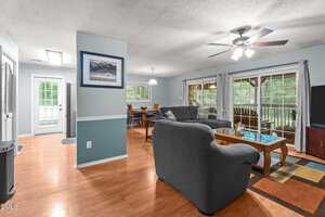 A living room with wood floors, gray furniture, a ceiling fan, and a large windowed door leading to a covered porch. A dining area and kitchen with bright lighting are visible in the background.