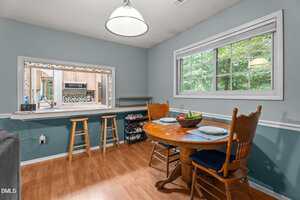 A small dining area with a round wooden table set for two, two wooden chairs, a window showing greenery outside, light blue walls, wood flooring, and a pass-through window to a kitchen with barstools.