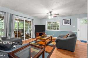 A living room with light blue walls, wooden floors, a ceiling fan with lights, gray couches, a wooden coffee table, a flat-screen TV, and large windows with curtains overlooking a covered patio.