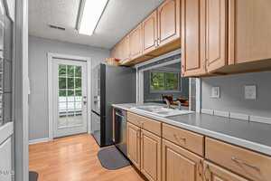 A kitchen with light wood cabinets, white tile countertops, stainless steel appliances, and wood flooring. There is a double sink, a window opening to the dining area, and a glass door leading outside.