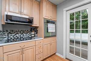 A kitchen with light wood cabinets, a black and white tile backsplash, an electric stovetop, built-in microwave and oven, utensils in containers, and a glass door leading to a deck with a white bench outside.