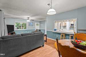 A living room with gray sofas faces a TV in the corner, adjacent to a dining area with a wooden table and chairs. A breakfast bar with stools separates the kitchen, and large windows let in natural light.