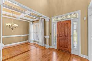 A spacious entryway with hardwood floors, a wooden front door with decorative glass panels, tall beige walls, and an adjacent dining area featuring a coffered ceiling, white trim, and a chandelier.
