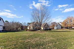 A single-story house with light-colored stone siding and a gable roof sits behind a large, leafless tree on a spacious, well-kept lawn under a blue sky with scattered clouds. Neighboring houses are visible on each side.