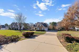 A wide concrete driveway leads to a two-story brick house with white trim and a three-car garage, surrounded by shrubs, trees, and a clear blue sky with scattered clouds.