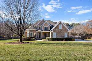 A two-story suburban house in Wake Forest with stone and siding exterior, gabled roof, and a manicured front yard featuring a bare tree, neatly trimmed bushes, and a wide driveway at 25 Sunflower Meadows Lane on a sunny day.
