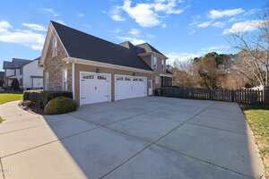 A two-story suburban house with stone and siding exterior, three-car garage with white doors, large driveway, and a fenced backyard under a blue sky with scattered clouds.