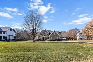 A two-story house with a stone and brick facade sits behind a large, leafless tree on a spacious lawn, with neighboring houses and trees visible on both sides under a blue sky with scattered clouds.