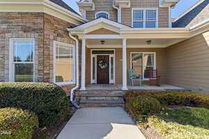 A front porch of a house with two columns, a dark wooden front door with a wreath, a blue rocking chair to the right, stone and siding exterior walls, and trimmed bushes along the walkway.