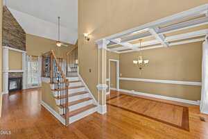 A spacious interior features a wooden staircase with iron railings, a coffered ceiling in the dining area, hardwood floors, beige walls, white trim, and a stone fireplace in the background.