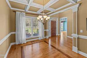A dining room with hardwood floors, beige walls, white ceiling beams, a central chandelier, large windows with curtains, and a wooden front door; an adjacent room with blue walls is visible.