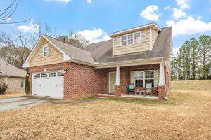 A two-story brick house with tan shingles, a double garage, a covered front porch with two chairs and a small table, and a brown lawn. Pine trees and a blue sky with clouds are in the background.