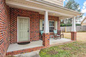 A brick house with a small covered front porch featuring two chairs, a small table, white columns, a pale green front door, and a window. The yard has grass and a wooden fence in the background.