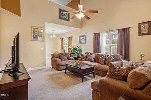 A living room with brown sofas, a coffee table, an armchair, and a TV on a stand. Large windows with curtains let in natural light. Ceiling fans and framed wall art are visible, and a dining area is in the background.