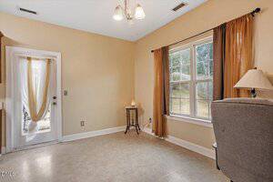 A small, empty room with beige walls and flooring, a window with brown curtains, a glass door with sheer curtains, a grey armchair, a lamp, and a small table holding a decorative item.
