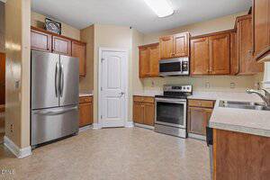 A kitchen with stainless steel appliances, including a refrigerator and electric stove, wooden cabinets, light countertops, beige walls, a white door, and tile flooring under fluorescent lighting.