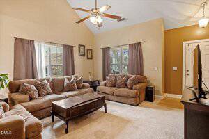A living room with two brown sofas, a coffee table, a TV on a stand, beige carpet, and taupe walls. Two windows with brown curtains let in natural light. Ceiling fan and hanging light fixture are visible.