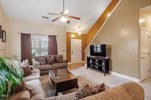 A living room with beige carpeting, tan and brown walls, a ceiling fan, large windows with curtains, a sectional sofa, a coffee table, a flat-screen TV on a stand, and a plant in the corner.
