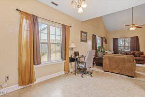 A home office area with a desk, office chair, and lamp near a window with brown curtains. In the background, a living room with a brown sofa set, a ceiling fan, and large windows is visible.