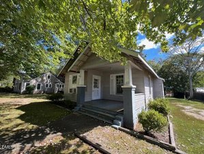 A single-story house at 1125 Beacon Avenue, Henderson, with light gray siding, a covered front porch supported by columns, and two white doors. The grassy yard features shrubs and is shaded by large leafy trees. Another house is in the background.