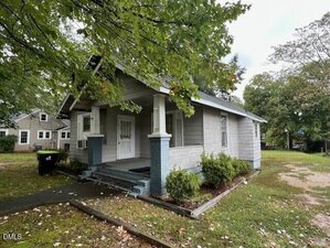 A small, single-story gray house with a covered front porch, white trim, two white columns with blue bases, and surrounded by green trees and grass. A sidewalk leads to the front steps.