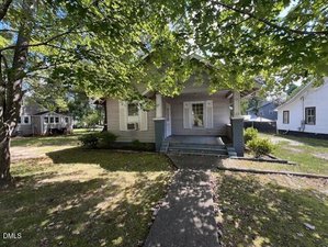 A single-story house with a covered front porch and white shutters, partially shaded by large trees. A concrete pathway leads to the front steps. The yard is grassy with scattered leaves. Neighboring houses are visible.