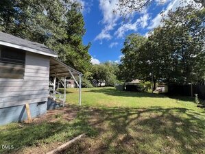 A grassy backyard with scattered trees, partial shade, and a small covered porch attached to a gray house on the left. The sky is mostly clear with some clouds. Other houses are visible in the background.