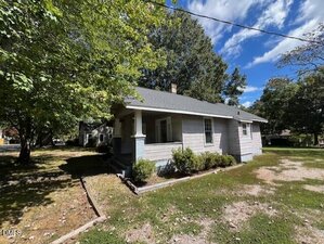 Single-story house with light gray siding, a covered front porch with columns, and a gable roof. Surrounded by trees, the yard is mostly grass with some bare patches and a dirt driveway. The sky is partly cloudy.