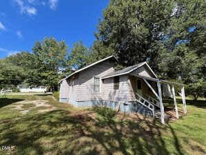 Single-story gray house with white trim and a covered front porch, set on a grassy lot with trees in the background on a clear, sunny day. Dirt driveway is visible to the left of the house.